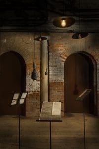 In front of brick arches, three metal stands hold printed pages while shaded bulbs and old-style microphones hang from the ceiling.
